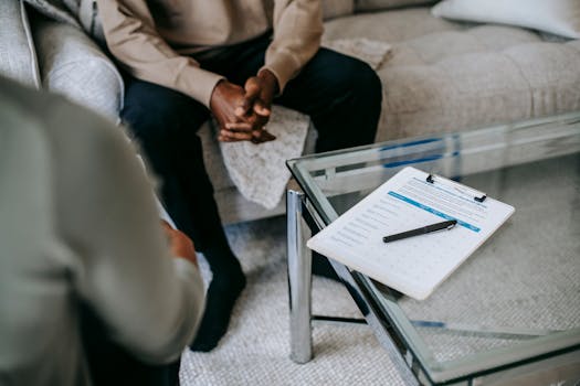 From above of unrecognizable ethnic male patient with clasped hands sitting near anonymous psychologist in office with glass table and clipboard with notes